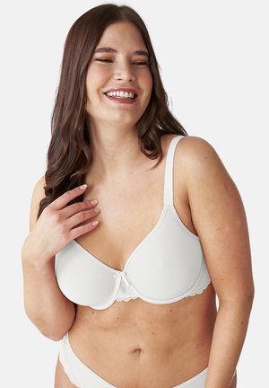 Woman with long brown hair smiling, wearing a white bra and matching underwear against a plain light background.