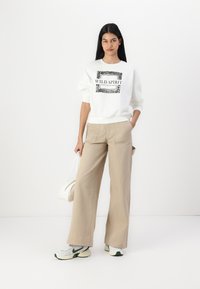 Witte sweatshirt met 'WILD SPIRIT' opdruk, gecombineerd met beige wijde broek. Model houdt een kleine witte handtas vast en draagt grijze sneakers.