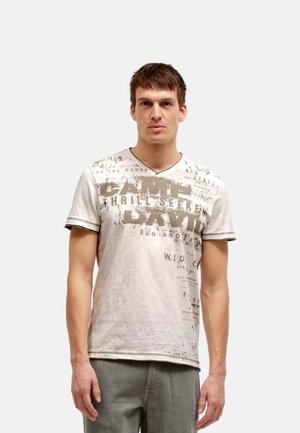 Mann trägt ein hellbeiges T-Shirt mit kurzem Arm und Grafikmotiv sowie graue Hose, steht vor einem einfarbigen hellen Hintergrund und blickt nach vorne.