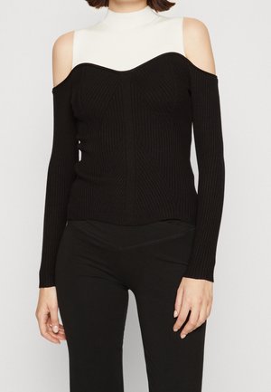 Femme portant un pull noir à épaules dénudées côtelé, superposé à un top blanc sans manches à col montant, avec un pantalon noir.