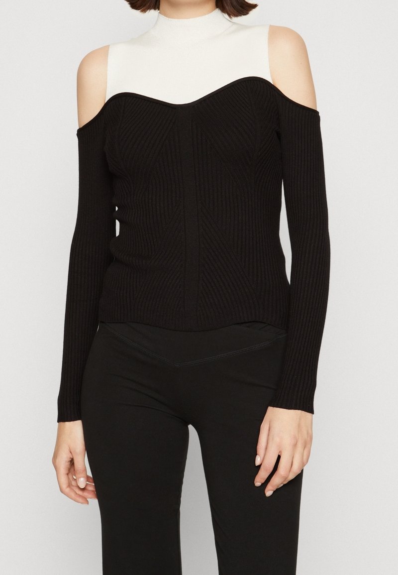 Femme portant un pull noir à épaules dénudées côtelé, superposé à un top blanc sans manches à col montant, avec un pantalon noir.