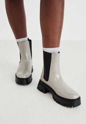 Personne portant des bottines Chelsea beiges brillantes et noires à semelles épaisses noires, avec des chaussettes blanches, sur un fond clair uni.