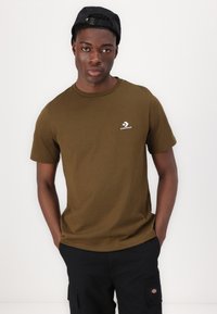 Camiseta de algodón verde oliva con cuello redondo y mangas cortas. Presenta un pequeño logo blanco de Converse en el lado izquierdo del pecho. Pantalones cortos cargo negros.