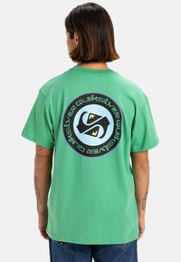 Camiseta de algodón verde con un gran gráfico circular en la parte posterior que presenta un logo en azul claro y negro, acompañado de un texto de contraste a su alrededor.