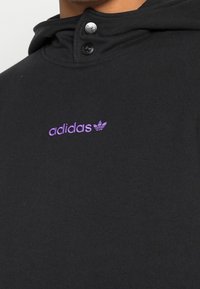 Närbild av svart hoodie med lila Adidas-logotyp broderad på bröstet och två svarta tryckknappar vid halslinningen.