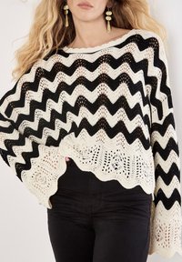 Schwarz-weißes Zickzackmuster-Poncho, gestrickt mit einem gewellten Saum und offener Webstruktur. Kombiniert mit schwarzen, schmalen Hosen.