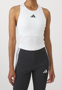 Femme portant un débardeur blanc de sport et un legging noir avec des rayures blanches et des logos, debout devant un fond uni.