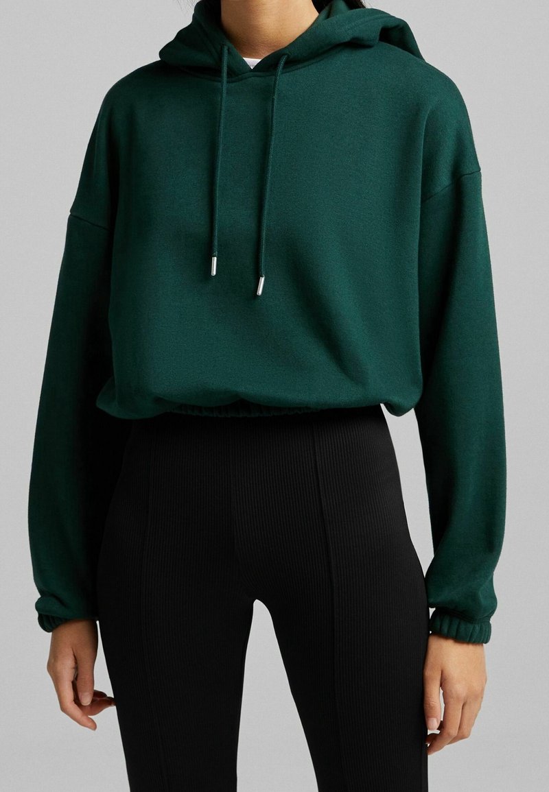 Personne portant un sweat à capuche court vert foncé avec cordons et des leggings noirs taille haute côtelés sur un fond uni.