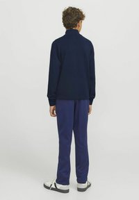 Pull navy avec un col montant, associé à un pantalon bleu assorti. Tissu texturé, debout avec les mains sur les côtés. Chaussures décontractées en noir et blanc.
