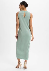 Mouwloze mintgroene jurk met een keyhole-detail op de rug, maxi-lengte en subtiele glinstering in de stof. Gecombineerd met metallic hakken.