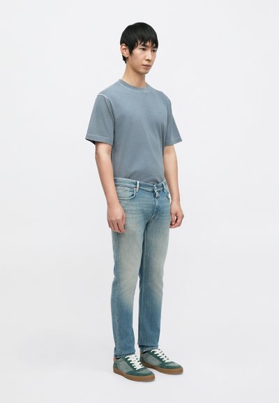 Μπλε βαμβακερό μπλουζάκι, ανοιχτό μπλε ξεθωριασμένο τζιν, πράσινα και γκρι αθλητικά παπούτσια με λευκά κορδόνια· πόζα όρθια μπροστά σε απλό φόντο.