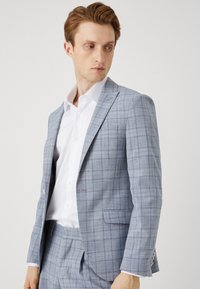 Costum cu carouri albastre deschise, cu o croială ajustată. Materialul pare a fi tesut, având o textură subtilă. Include o cămașă albă de birou dedesubt.