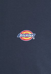 Tecido azul-marinho com um logotipo circular vermelho, amarelo e azul com "Dickies" no centro. Textura suave, design ajustado.
