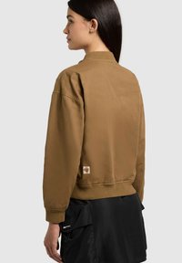 Woman in profile wearing a tan bomber jacket with ribbed cuffs and hem, paired with a black skirt featuring front pockets.