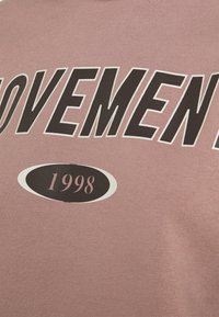 Różowa bawełniana koszulka z wyraźnym czarnym napisem "RUCH" oraz rokiem "1998" w okrągłym wzorze. Miękka tekstura, swobodny fason.