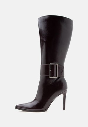 Botte haute en cuir marron foncé à talon haut, bout pointu, avec une large sangle dotée d'une grande boucle métallique rectangulaire autour de la cheville.
