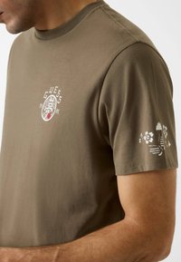 Grünes Baumwoll-T-Shirt mit kurzen Ärmeln, mittig mit weißem Logo und floralem Muster auf dem Ärmel. Glatte Textur.