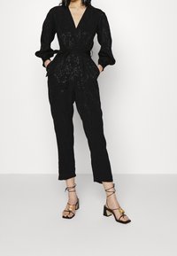 Svart jumpsuit med v-ringning, puffärmar och knytmidja. Har en intrikat blommönster. Matchad med svarta sandaletter med remmar och klack.