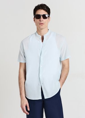 Hellblaues kurzärmeliges Hemd mit Stehkragen, strukturierter Stoff und lockerer Passform. Kombiniert mit dunkler Hose und Sonnenbrille.