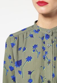 Olivově zelená blůza s modrým květinovým potiskem, s pevně stojacím límcem, zapínáním na knoflíky a plisovanými detaily na přední části.