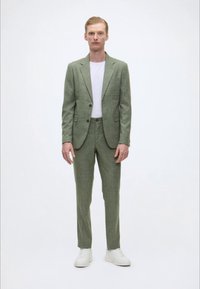 Hellegrüner Anzug aus strukturiertem Stoff. Maßgeschneiderte Blazer mit zwei Knöpfen, passende Hose. Trägt man über einem weißen T-Shirt, kombiniert mit weißen Turnschuhen.