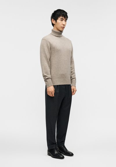 Πουλόβερ ζιβάγκο μπεζ, από μάλλινο υλικό, με ριμπ μανσέτες, σε συνδυασμό με σκούρα παντελόνια με ρίγα, και μαύρα παπούτσια με κορδόνια. Στέκεται μπροστά σε ουδέτερο φόντο.