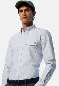 Camisa de botones a rayas en azul claro y blanco, con un bolsillo en el pecho y un pequeño parche con un logo. Se lleva con un cinturón oscuro y pantalones grises.