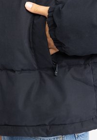 Čierna prešívaná bunda s vreckom na zips, rebrovanými manžetami a jemnou textúrou. Tkanina sa zdá byť odolná a vodoodpudivá.