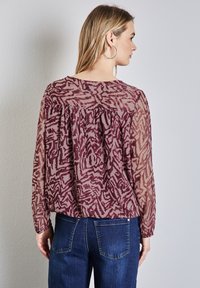 Transparante blouse met lange mouwen in bordeaux met een abstract patroon, voorzien van een ronde halslijn en een geraffineerd detail aan de achterkant, gedragen met donkere spijkerbroeken.