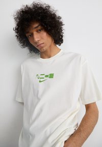 Hvid bomulds-T-shirt med grøn tekst og logo på forsiden. Korte ærmer, afslappet pasform og glat tekstur.