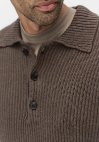 Brun stickad tröja med ribbad textur, med en vikbar krage och tre svarta knappar framtill. Nära vy som framhäver materialet.