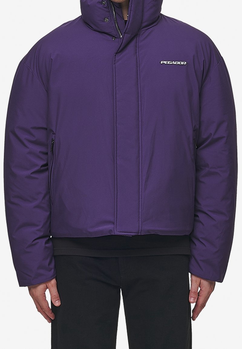 Veste isolante violette avec un col haut, fermeture éclair devant, poches latérales et coupe courte. Présente un logo sur la poitrine. Texture lisse.