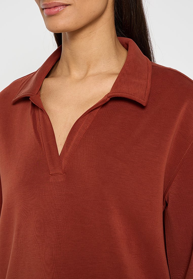 Haut couleur rouille avec un large col, présentant un design en V et une texture douce. Coupe légèrement décontractée, adaptée à une tenue décontractée.