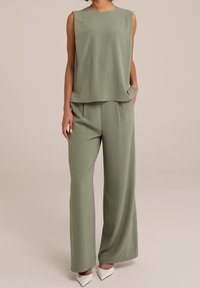 Kobieta ubrana w luźną, bezrękawnikową oliwkową bluzkę i pasujące szerokie spodnie, na białych czółenkach, stoi z jedną ręką w kieszeni.