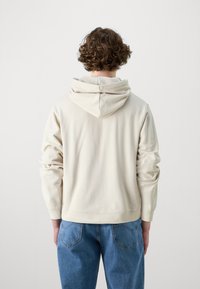 Beige hoodie i mjukt material med dragsko i huvan, avslappnad passform och ribbade mudd. Kombinerad med blå denimjeans.