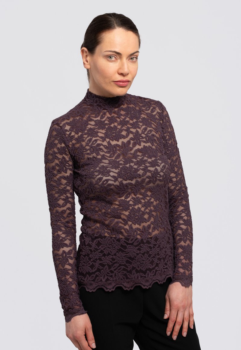 Top de encaje en púrpura oscuro con mangas largas y cuello alto. Presenta patrones florales y un dobladillo a rayas. Se lleva con pantalones negros.
