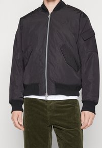 Svart bomberjacka med dragkedja fram, ribbade manschetter och nederkant, sidofickor och en axelficka. Har en logotyp på bröstet.