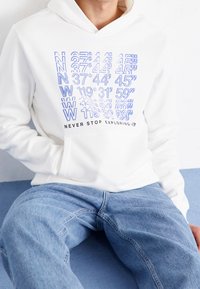 Sudadera blanca con coordenadas geográficas impresas en azul y el texto "NUNCA DEJES DE EXPLORAR". Combinada con jeans de mezclilla azul claro.