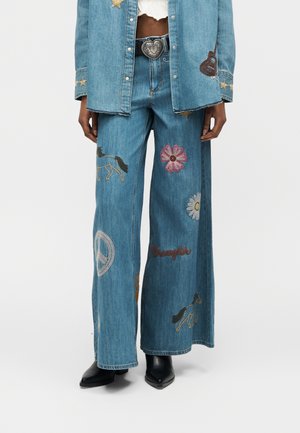 Jean en denim à jambes larges avec broderies de cheval, fleurs, symbole de paix et texte "Wrangler", assorti à une veste en denim et des bottes noires.