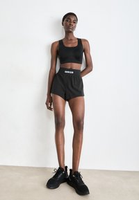 Cropped top sportivo nero e pantaloni corti a vita alta con banda in vita di Calvin Klein. Il modello indossa sneakers nere, in piedi su uno sfondo neutro.