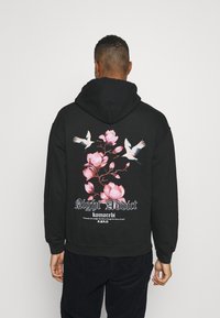 Sudadera negra con un diseño floral que presenta flores rosas y pájaros blancos en la parte posterior, con el texto "Night Addict" y "komorebi."