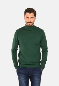 Maglione verde a collo alto realizzato in tessuto morbido, con un design aderente e polsini a coste, abbinato a jeans di denim blu.