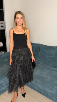 Black sleeveless top paired with a tiered black tulle skirt featuring a sparkly texture. Black pointed-toe flats. Black clutch in hand.