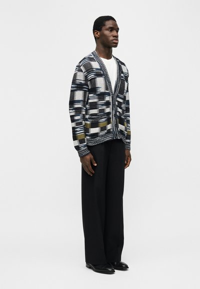 Άνδρας με μαύρο παντελόνι φαρδιάς γραμμής, μαύρη και λευκή ζακέτα με σχέδιο και ελαιώδεις αποχρώσεις, λευκό πουκάμισο και μαύρα παπούτσια, στέκεται μπροστά σε απλό φόντο.