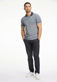 Polo grigio con colletto blu navy e dettagli a righe, abbinato a pantaloni scuri e sneakers bianche, mostrato su un modello maschile su uno sfondo neutro.