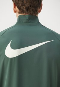 Nike-takki tummanvihreästä kankaasta, jossa on korkea kaulus, sileä rakenne ja suuri valkoinen swoosh-logo takana.