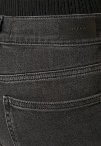 Jeans de mezclilla negra con un acabado texturizado y ligeramente desgastado, con costuras en contraste. Incluye un parche de cuero con el logo en la cintura.
