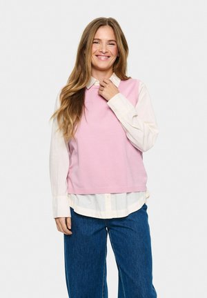 Un gilet en tricot rose clair superposé sur une chemise blanche à boutons. Un jean en denim bleu ample. Design simple avec ourlet et encolure côtelés.