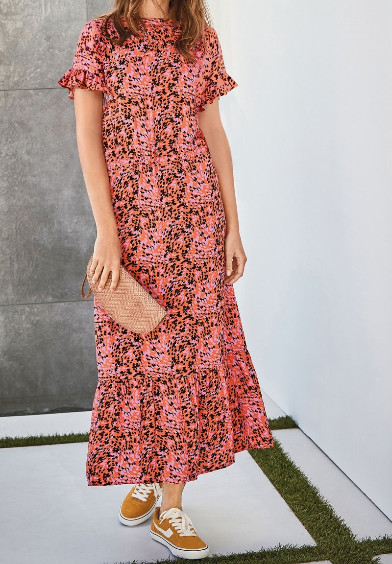 Femme portant une robe midi rose et noire à motifs avec des manches à volants, tenant une pochette tissée, et portant des baskets jaune moutarde.