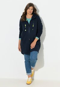 Námořnicky modrá mikina na zip s texturovaným povrchem a neonovými šňůrkami. Kombinováno s vyhrnutými džíny a žlutými teniskami.